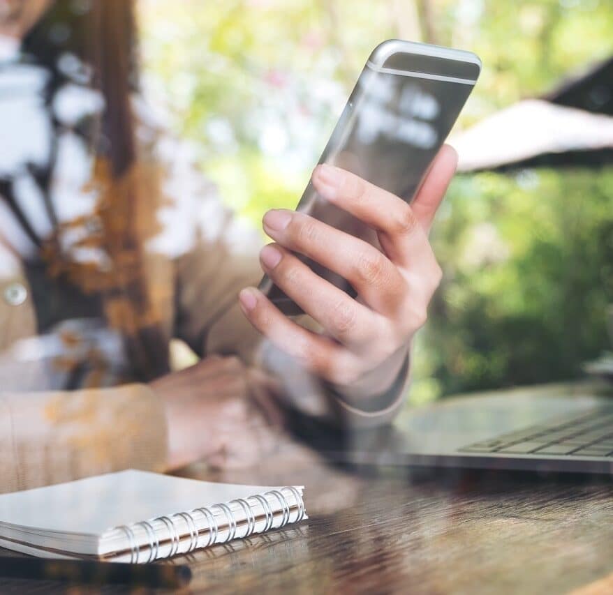 Closeup image of woman's hand holding and looking at smart phone while using laptop in cafe